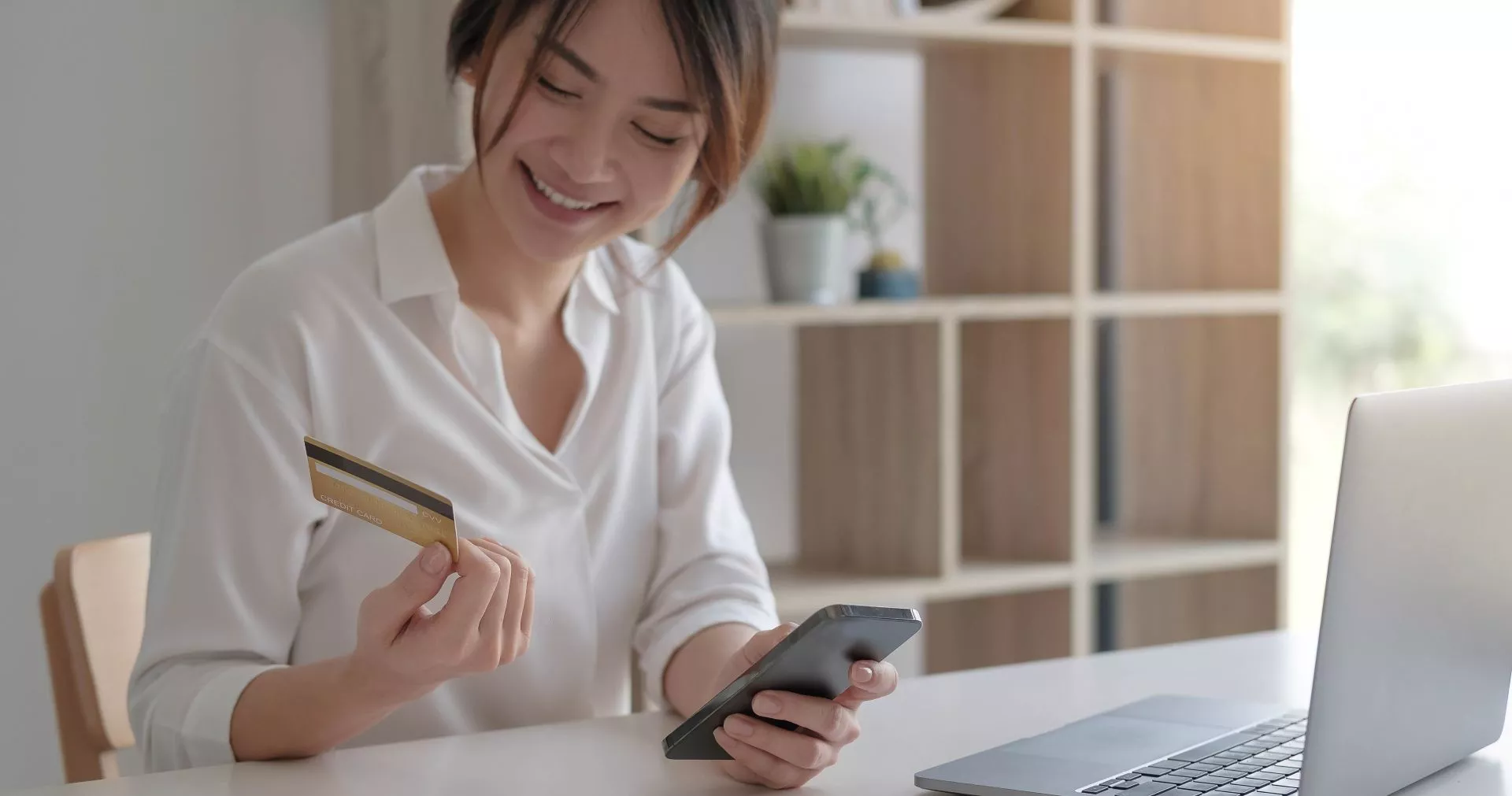 202602021798.webp 女性在室內桌前微笑看著手機,手中拿著信用卡,桌上放有筆電,呈現線上購物或數位支付情境。